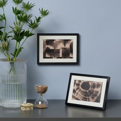 Framed photo & abstract art against wall. Glass vase with plants & hourglass.