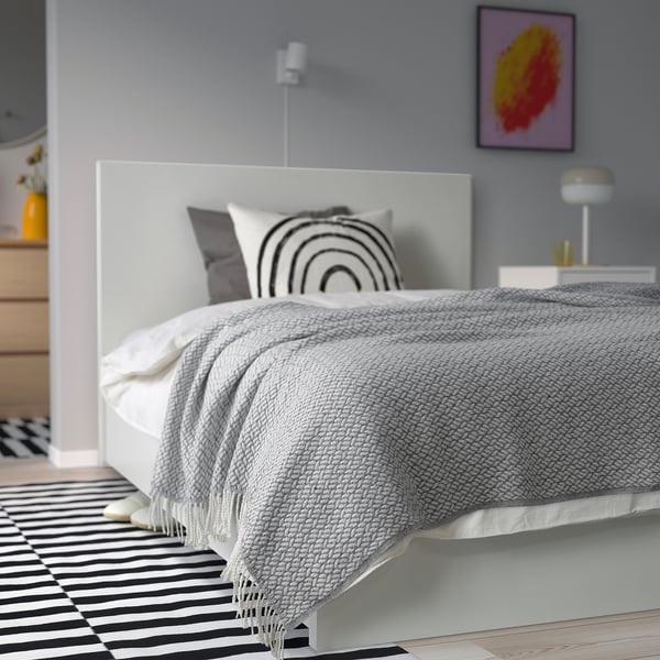 Grey MAJINGER throw on white bed, striped rug, minimalist decor.
