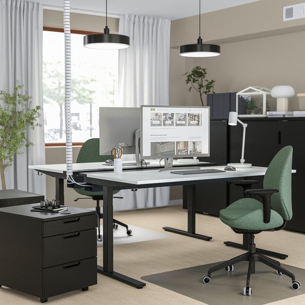Modern office setup with MITTZON black height-adjustable desk, green chairs, dual monitors, plants, and minimalist decor.