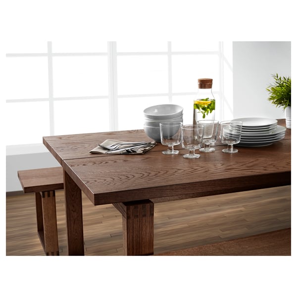 Modern wooden dining table set with clear glasses, white dishes, grey napkins, and a bottle with greens.