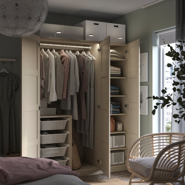 Beige PAX wardrobe with open doors, clothes, and shelves. Hanging garments, towels, baskets. Near window with plant, wicker chair.