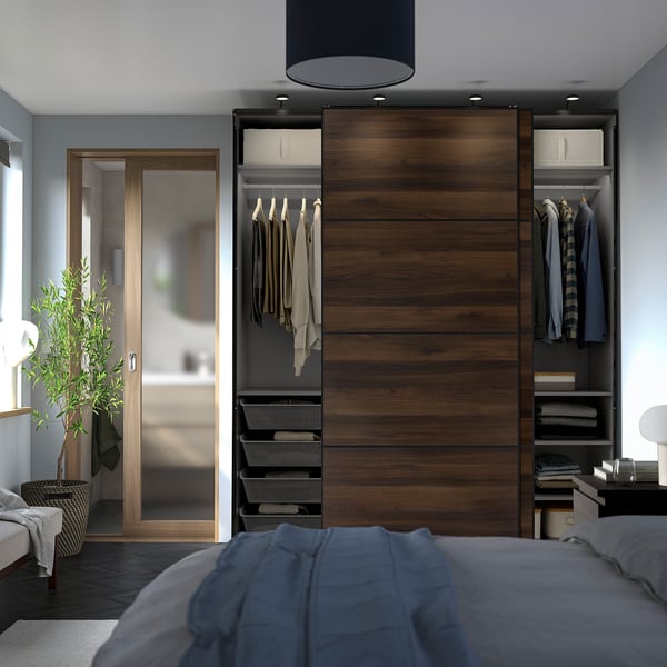 Modern bedroom with large wooden PAX wardrobe featuring sliding doors, open shelves, and hanging clothes. Adjacent glass-doored closet.