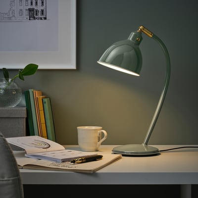 A green RÖDFLIK desk lamp on workspace, shiny enamel, brass accents, adjustable shade, illuminating mug, notebooks, vase.