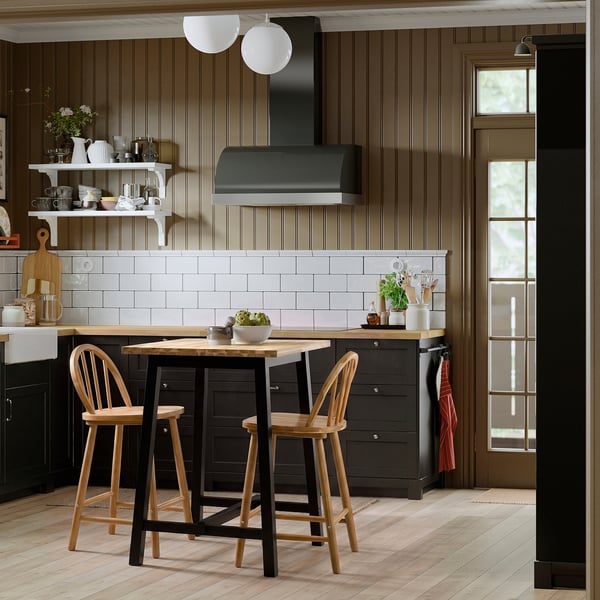 SKOGSTA bar table with black legs and acacia top, wooden stools, dark wood panelling, white subway tiles, stainless steel hood, window.