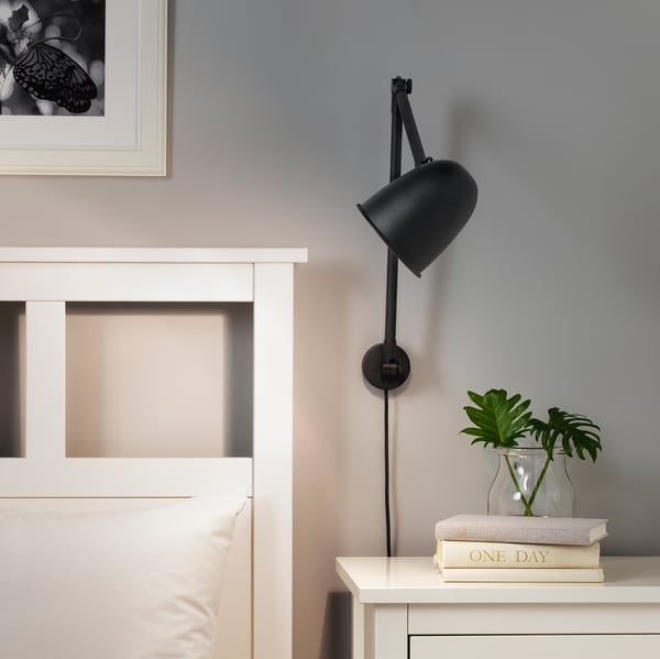 Black adjustable wall lamp above white nightstand with plant and book.