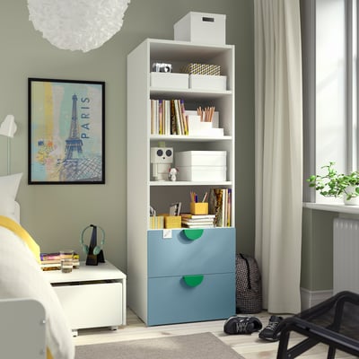 SMÅSTAD bookcase in light blue with green accents, three shelves, two drawers, near paris-themed artwork, bed, and black chair.