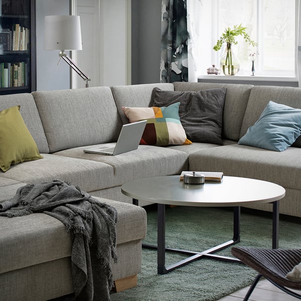 Modern living room with grey SÖRVALLEN sofa, colourful pillows, laptop, grey throw, coffee table, lamp, bookshelf, curtains, and potted plants.