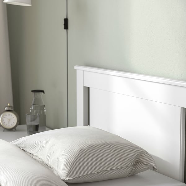 Bedroom scene with white bed frame, striped pillows, glass pitcher, and clock on bedside table.