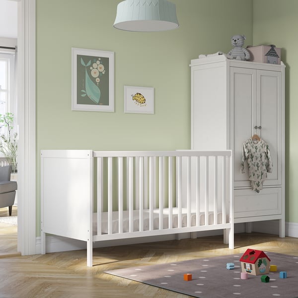 SUNDVIK white baby crib with adjustable base, near wardrobe, toys & rug.