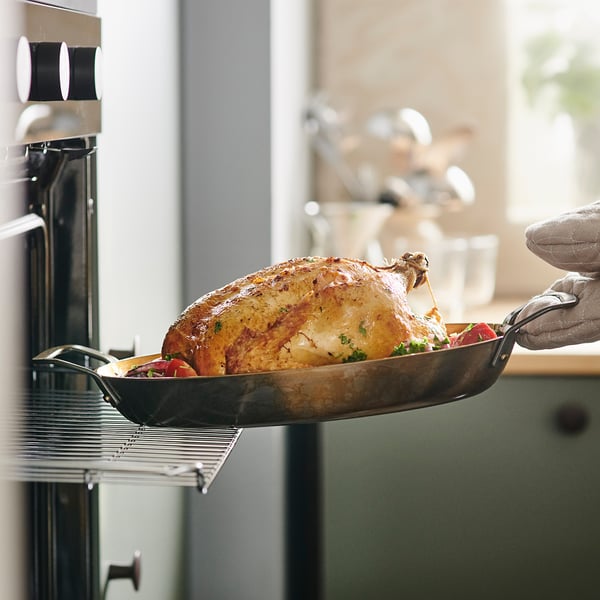 A person holding a roasted chicken in a dark roasting pan, showing even browning and heat retention for delicious dinners.
