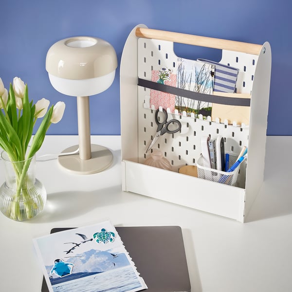 A white desk organiser with a handle, featuring hooks with various items, standing next to a lamp and flowers.