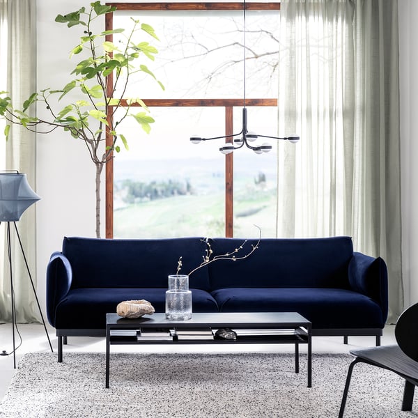 Modern living room with navy blue ÄPPLARYD sofa, glass coffee table, and large windows with curtains. A stylish and comfortable lounge space.