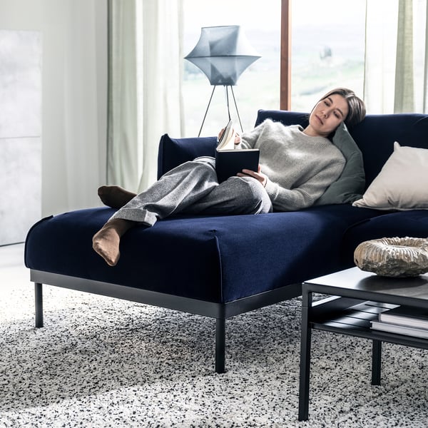 A person relaxes on a blue ÄPPLARYD chaise sofa, reading a book beside a modern lamp.