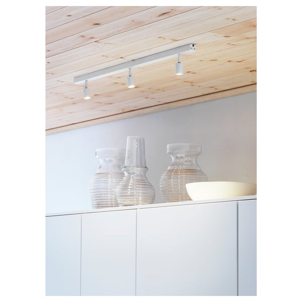 Modern ceiling track lights with three adjustable spots illuminate clear, cylindrical vases on a sleek white cabinet.