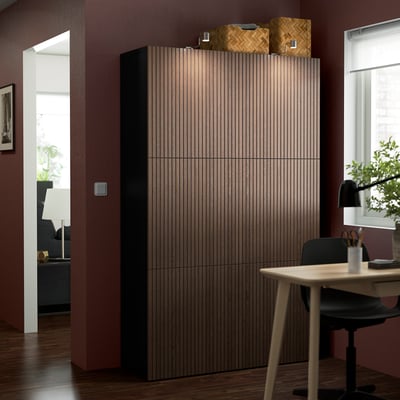 Large light brown BESTÅ storage unit with vertical lines, adjustable shelves, and wooden doors, standing tall next to a table and chair.