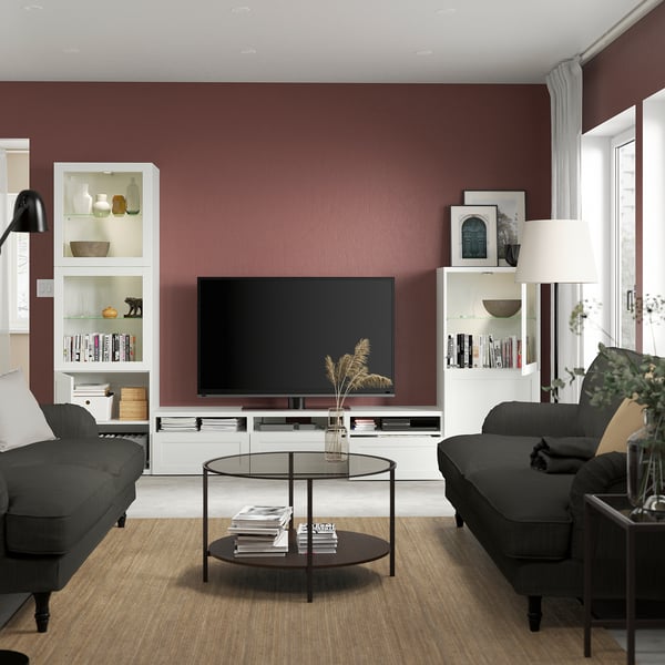 Modern living room with BESTÅ tv unit and flat-screen tv on maroon wall. Grey sofas flank a glass coffee table with decor and books.
