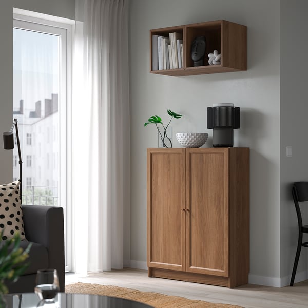 Bright room with brown BILLY bookcase. Holds lamp, bowl, plant. Between chair and window, against grey wall.