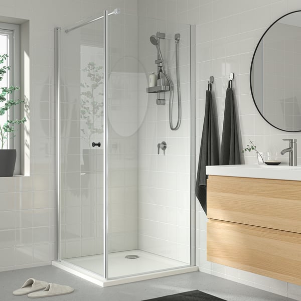 Modern bathroom with white tiles, glass shower, dark towels, wooden vanity, chrome fixtures, round mirror, plant, slippers by shower.