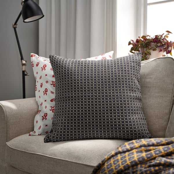A grey BRUNNYCKLAR cushion on a sofa, larger than the adjacent lamp and flowers, with a subtle textured pattern.