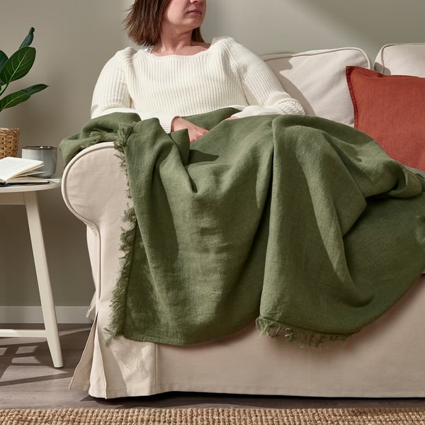 Person sitting on cream-colored couch, wrapped in green throw blanket with fringe, next to side table.