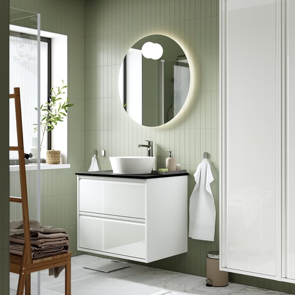 Modern bathroom with white EKOLN sink vanity, round mirror, and green tiles. Sink small compared to vanity. Towels and plant nearby.