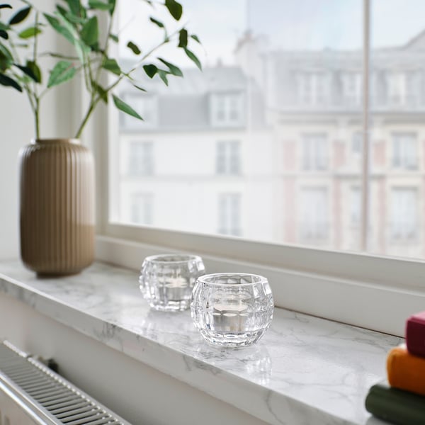 Two clear FRÖJDA glass candle holders on a marble windowsill, smaller than a vase with greenery.
