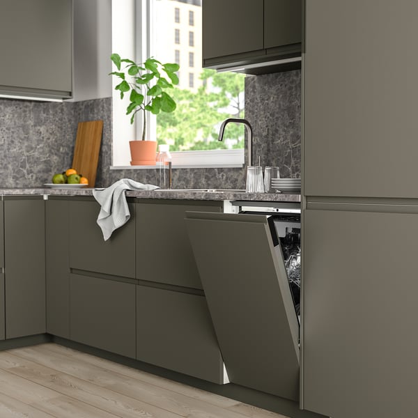 Modern kitchen with dark grey FRÖSLUNDA dishwasher open under granite countertop, next to plant and cutting board, sinks ahead, window far.