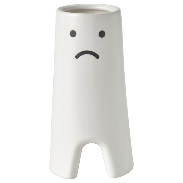A white, cylindrical vase named FRÖVÄXT with a sad expressive face design. Its made of stoneware and has an opening at the bottom.