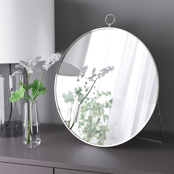 A round mirror stands on a surface reflecting a vase with flowers; it can be hung or placed.