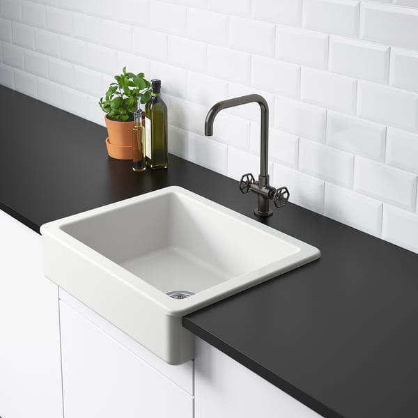 Modern kitchen sink with white bowl, dark countertop, matte black tap, white tiled wall, plants, and bottle.