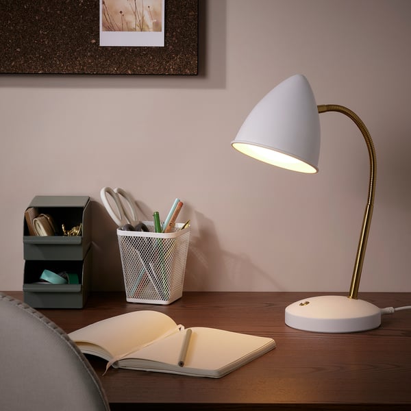 ISNÅLEN lamp in white with brass tube, flexible neck, illuminating desk with book and office supplies.