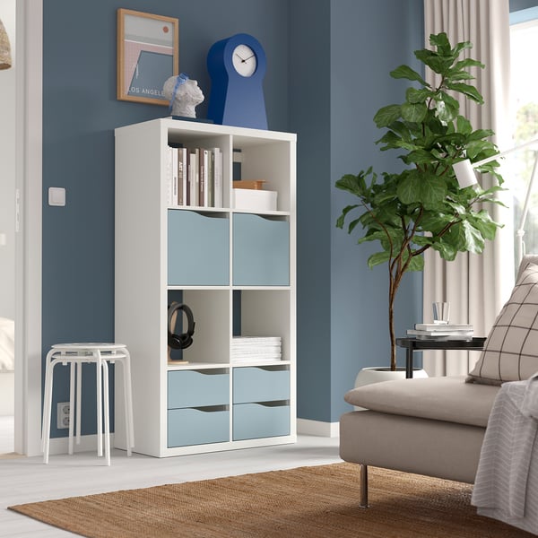 Modern living room with white KALLAX shelving, books, clock, figure, stool, beige couch, plant.