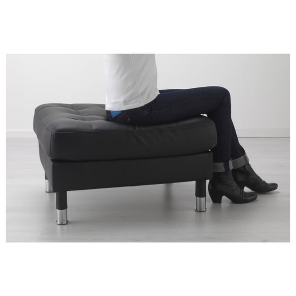 Person sitting with legs crossed on black modular landskröna footstool, featuring padded cushion and chrome legs.
