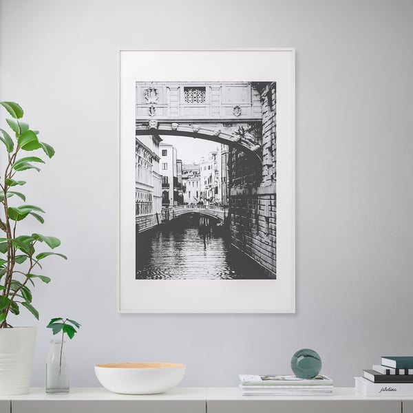 Framed black-and-white canal photo on wall. Shelf below with plant, bowl, vase, books, and decorative sphere.