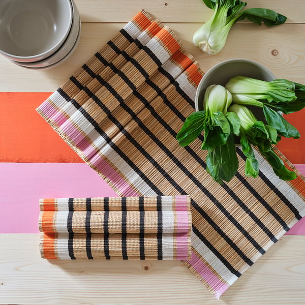 Colourful striped woven table runner on wood, with bowls and greens. Protects and decorates table; handmade with natural materials.