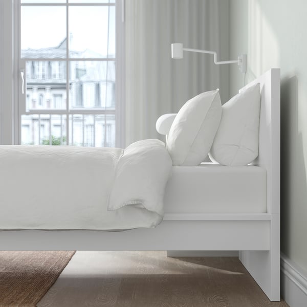White modern bed by window, pillows stacked, natural light.