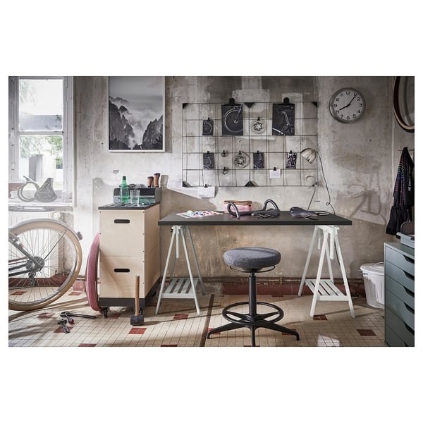 Industrial-style home office with MITTBACK black table, white legs, grey stool, concrete wall, clock, art, bike, and cabinet.