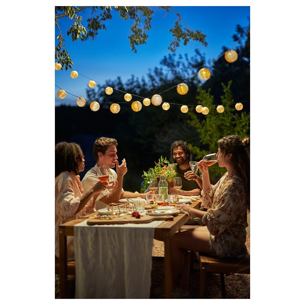 Four people enjoying dinner outdoors under hanging lanterns, seated at a rustic wooden table set with plates, glasses, and centrepieces.