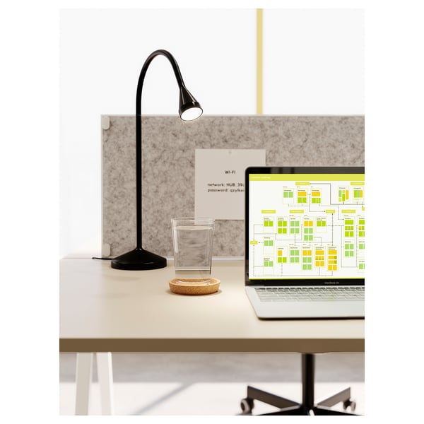 Black NÄVLINGE lamp lights workspace with macbook and network diagram, alongside a glass cup and cork coaster.