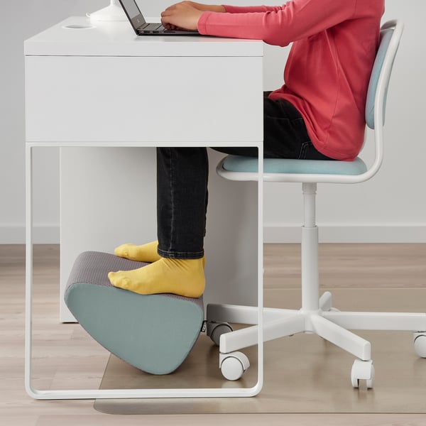 Person sitting at white desk, typing on laptop. Their feet rest on grey footstool, promoting better posture and blood circulation.