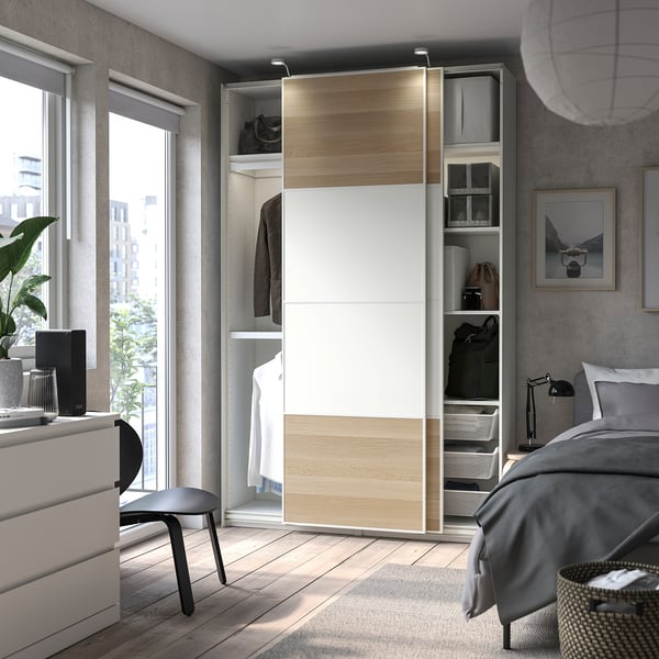 Modern bedroom with PAX wardrobe, sliding doors, grey bedding, open shelves, and black chair.