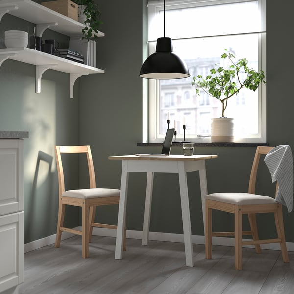 Modern minimalist kitchen corner with white table and wooden chairs. Black lamp, green plant, open shelves with dishes. Perfect for compact spaces.