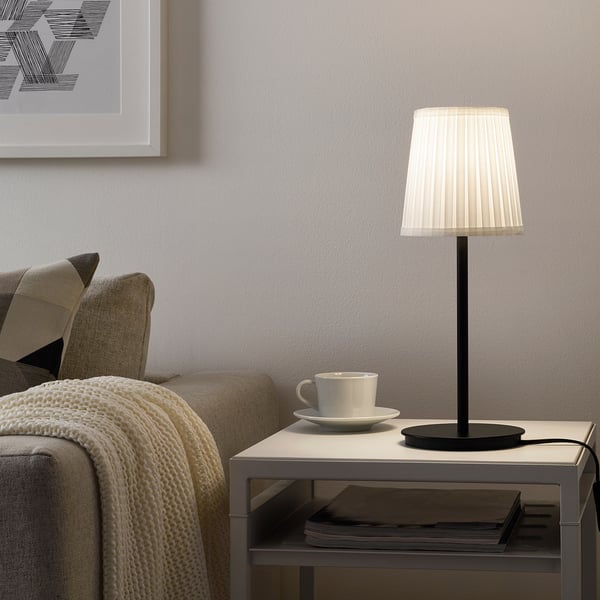 A cosy living room with SKAFTET lamp beside a cream sofa, knitted blanket, and magazines. Grey wall with a picture.