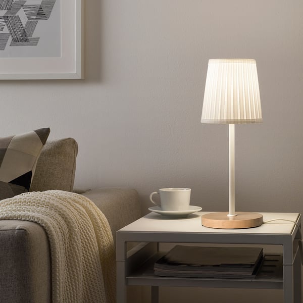 Small, round SKAFTET lamp on table, next to coffee mug, next to couch with blanket.