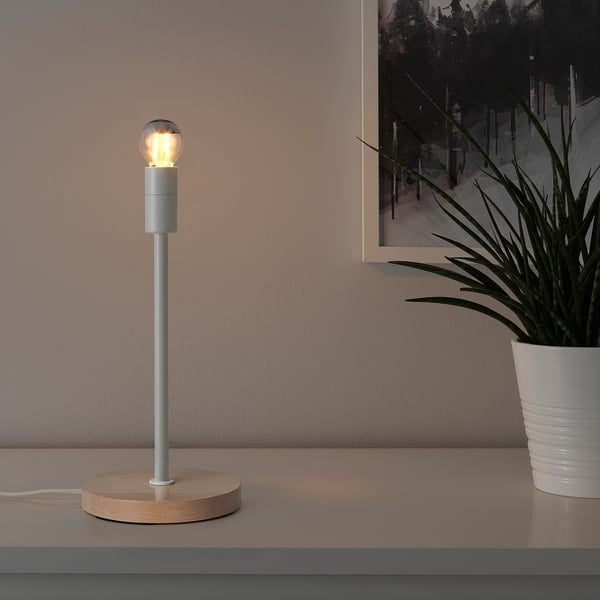 SKAFTET silver lamp on table, small, with light bulb on, next to white plant.