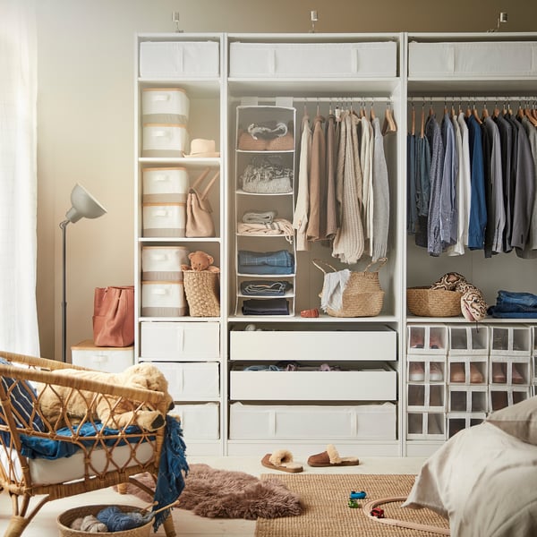 Well-organized closet featuring white SKUBB storage boxes, hanging clothes, woven baskets, and a wicker chair.