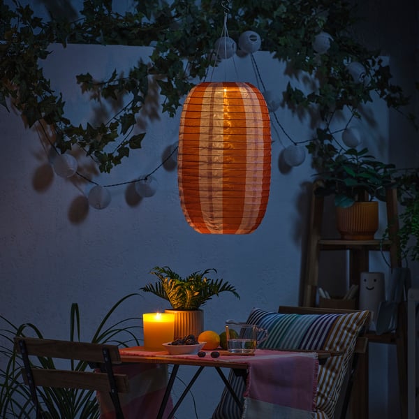 Cosy outdoor setting with a glowing SOLVINDEN solar lamp, candle, plants, and striped pillows.