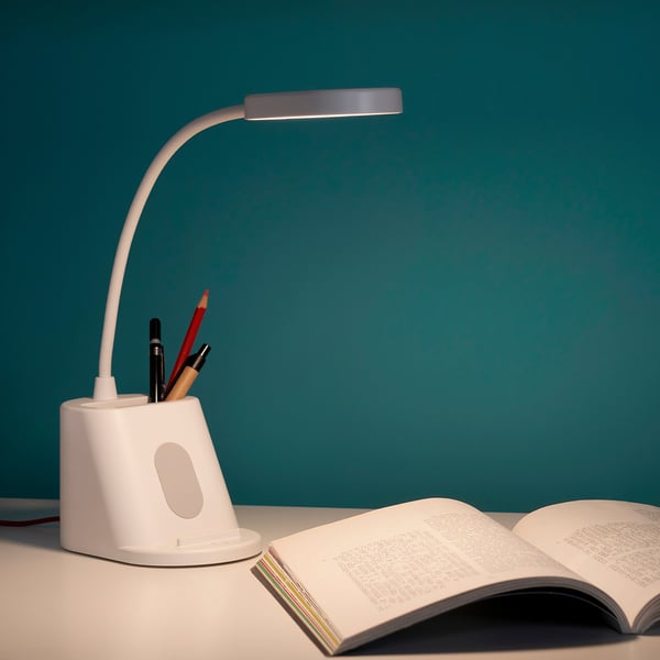 SPANNLAND lamp & holder in white on desk. Small, curved lamp illuminates open book. Holds pens, phone, charges via usb.