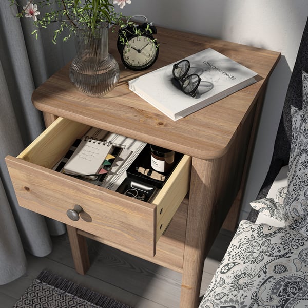 Wooden bedside table with open drawer showing items. Includes books, glasses, clock, and vase with flowers on top. Drawer organises small items.