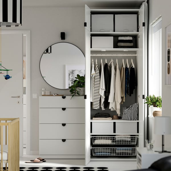 Open wardrobe with clothes, folded items in boxes. White drawers and mirror beside it. Modern nursery setup.